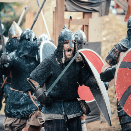 Schreiender Ritter stürmt mit weiteren Rittern aus einem Holztor. Dabei hält er ein Schild und Schwert und trägt ein Kettenhemd und einen Nasalhelm.